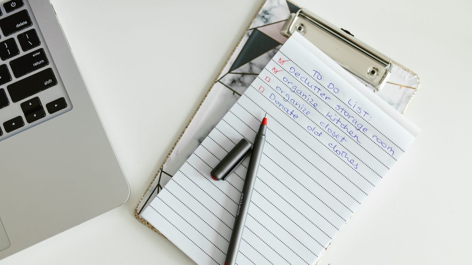Notepad checklist and pen on a clipboard beside a laptop on a white desk